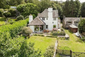 Una casa de dos pisos con un jardín amplio, un patio con mobiliario exterior y un camino de piedra que lleva a la casa.