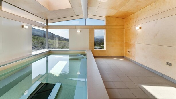 En modern inomhuspool med väggar av genomskinligt glas, träpanel och stora fönster som erbjuder utsikt över ett bergigt landskap.