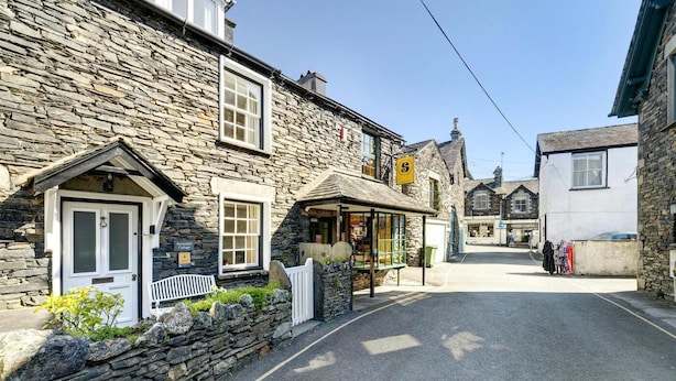 A stone building with a white door and windows, a small shop with a yellow sign, and a clear street. - 3 Bed in Ambleside (oc-lr121) (Ambleside)