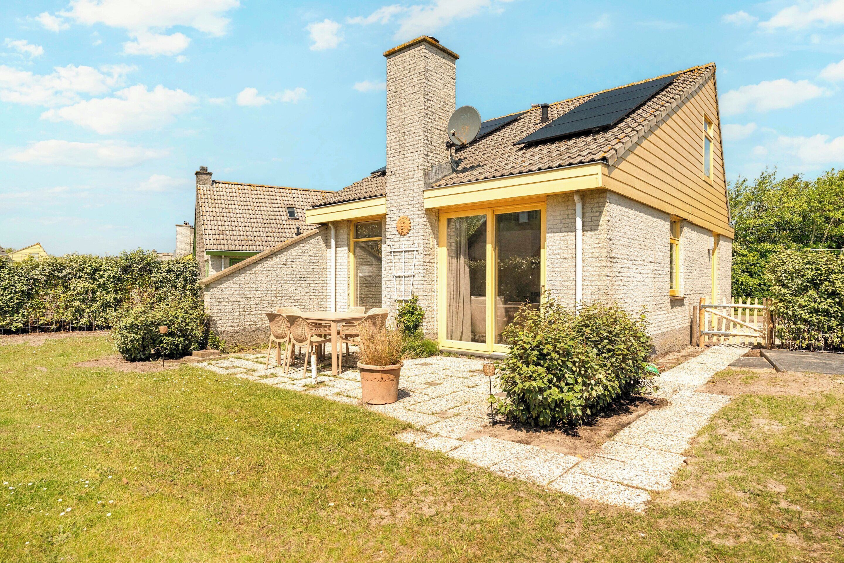 A house with a stone facade, a chimney, a solar panel, a patio with a table and chairs, and a garden.