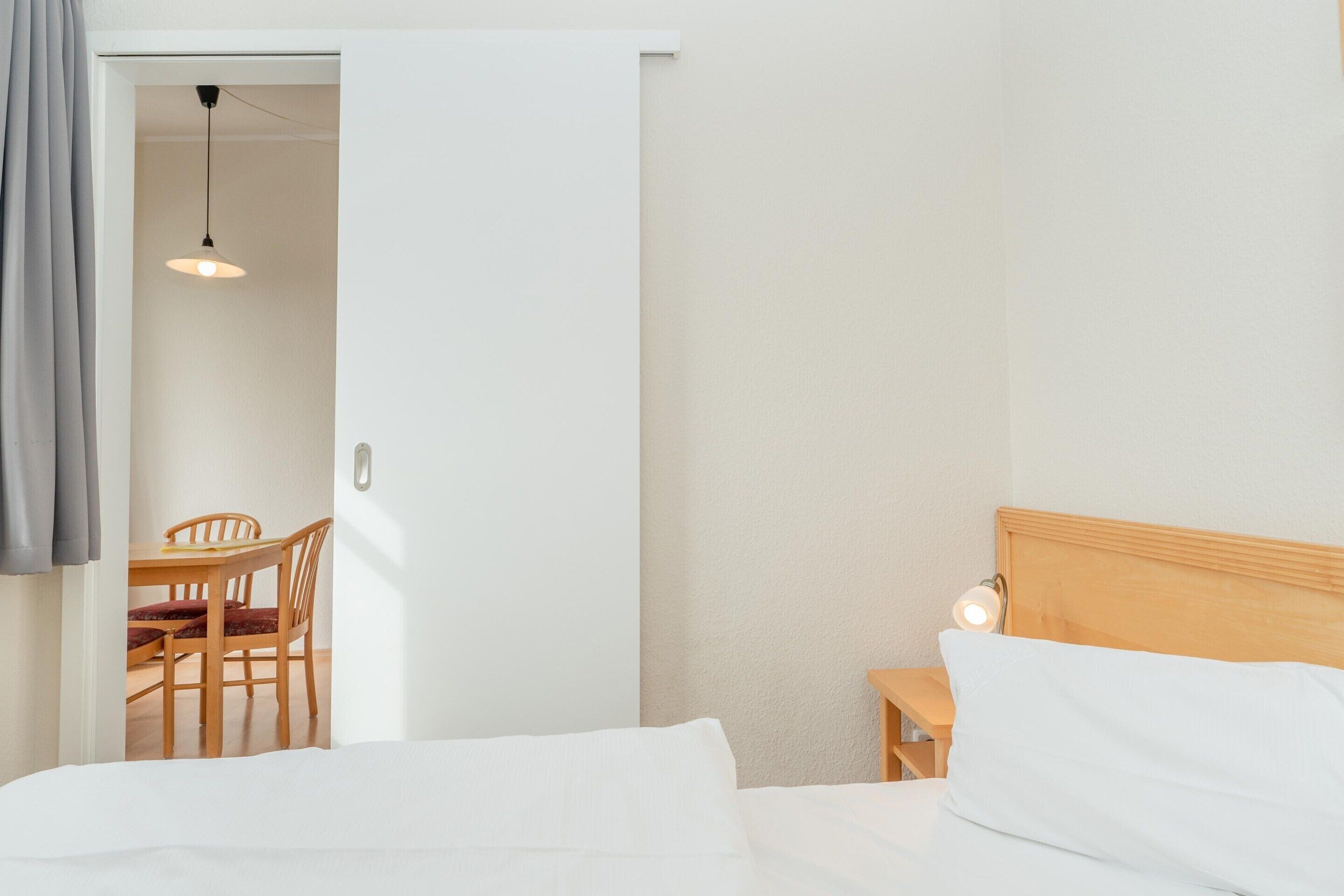 A bedroom with a bed, a bedside table with a lamp, a wooden headboard, and a door leading to a dining area with a table and chairs.