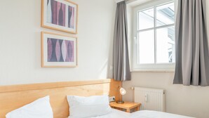 Hotellihuone, jossa on puinen sängynpääty, valkoisilla lakanoiden peitetty sänky, yöpöytä lampun kanssa ja kaksi kehystettyä kuvaa seinällä.