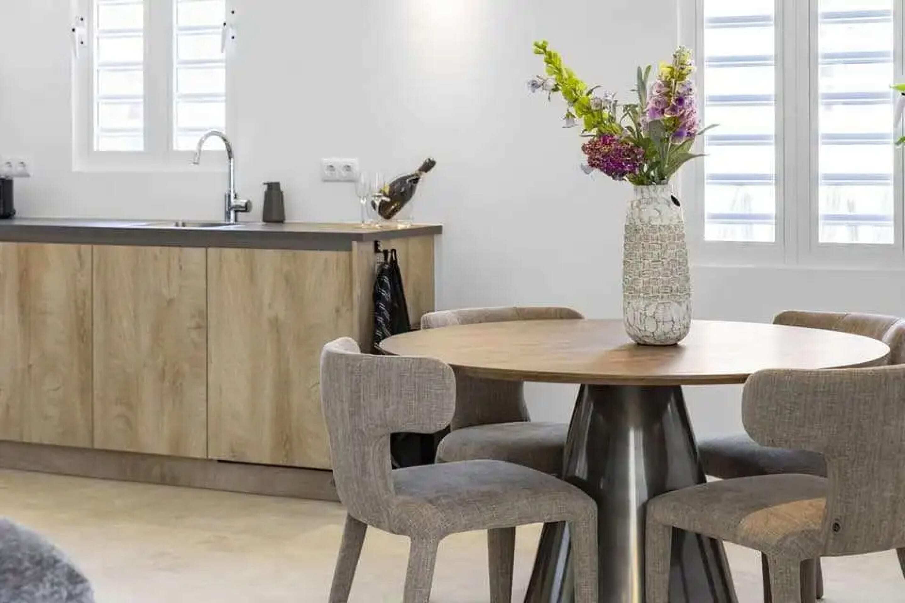 A modern kitchen with a wooden countertop, a round dining table with grey chairs, and a vase with flowers on the table.