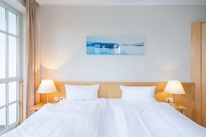 A hotel room with a bed, two bedside lamps, a window with curtains, and a painting on the wall. - Willkommen in Einer Modernen Ferienwohnung (Sellin)