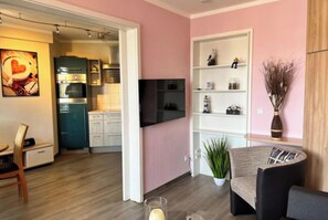 Ein modernes Wohnzimmer mit Fernseher, einer Couch, einem Esstisch und einer durchgehenden Tür erkennbaren Küchenzeile.