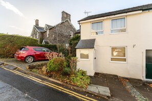 A residential street with a red car parked on the side, a white house with two windows, and a stone building in the background. - 2 Bed in Keswick (oc-lr147) (Keswick)