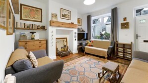 Ein gemütliches Wohnzimmer mit einer Couch, einem Sessel und einem kleinen Tisch. Es gibt Bücherregale, einen Spiegel und ein Fenster mit Vorhängen.