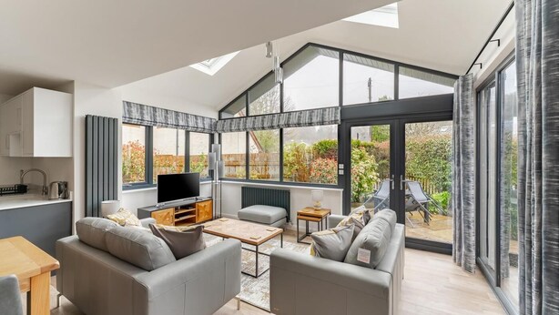 Ein modernes Wohnzimmer mit Sofa, Couchtisch und Fernseher, großen Fenstern und Blick ins Freie.