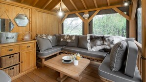 Een gezellig chaletinterieur met een houten plafond, een bank met een bont plaid, een bijzettafeltje met een bloemdecoratie, en een metalen container met de tekst 'BEER' op een houten kast.