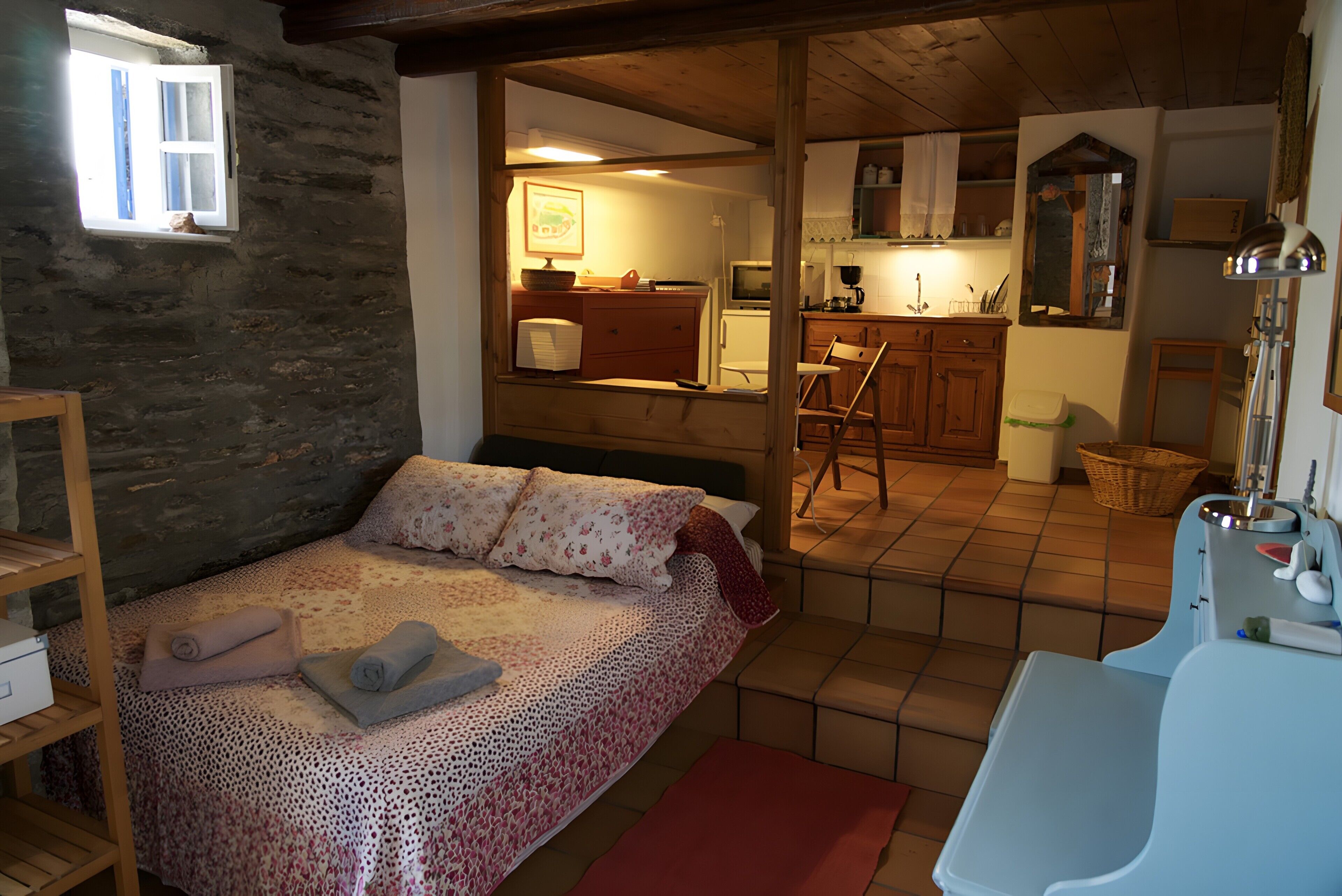 A bedroom with a bed, a small window, a kitchenette, and a bathroom with a sink and mirror.