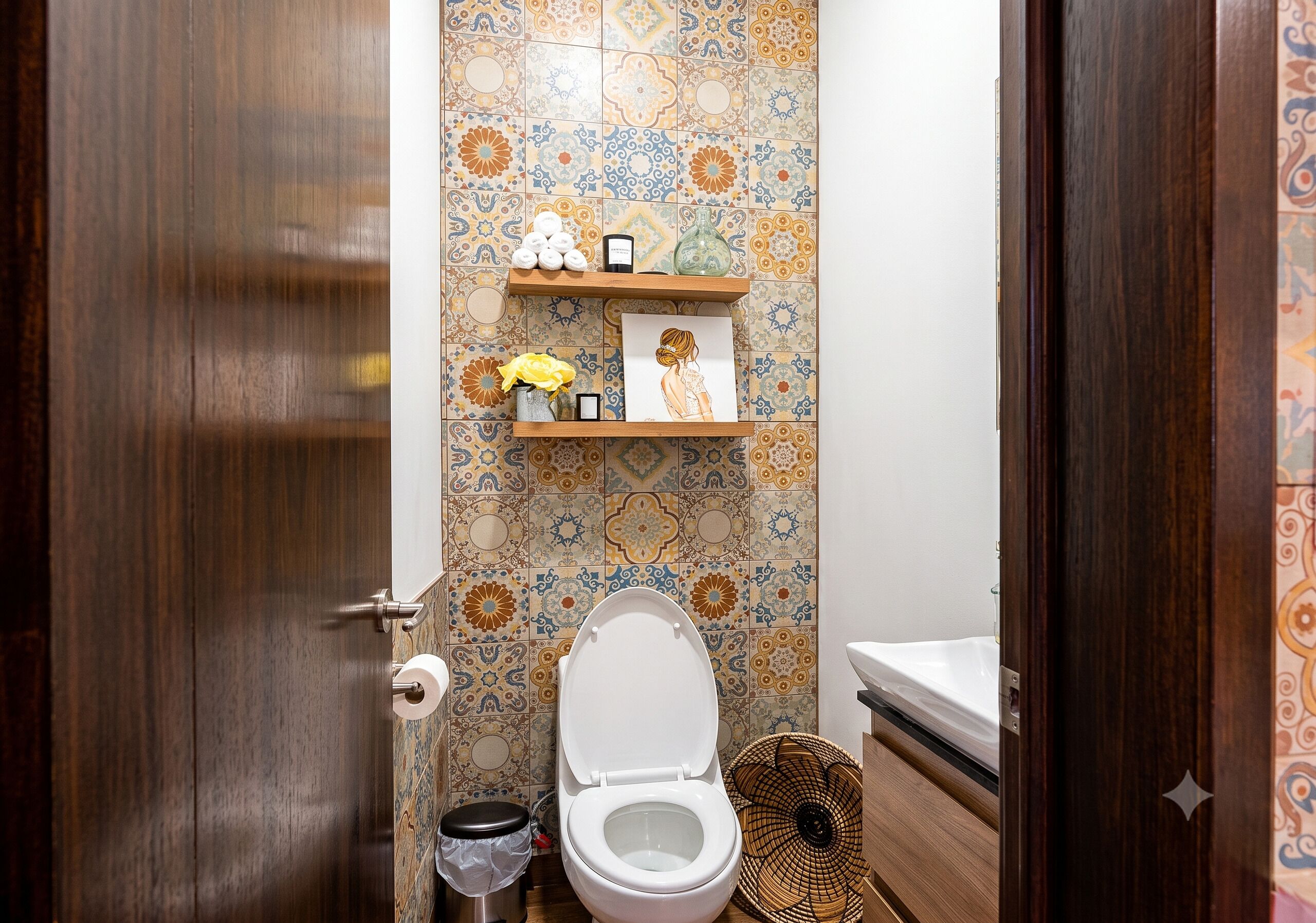 A bathroom with a toilet, a shelf with decorative items, and a patterned tile wall.