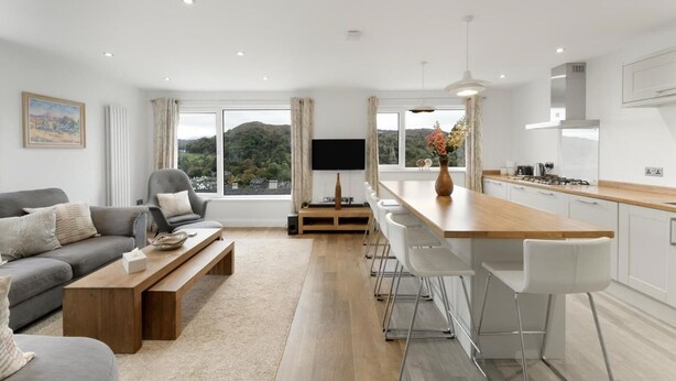 Un salon moderne avec un canapé, une table basse et un téléviseur, une cuisine avec un bar et de grandes fenêtres offrant une vue sur les collines.