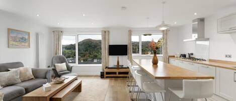 Ein modernes Wohnzimmer mit Sofa, Couchtisch und Fernseher, eine Küche mit Bar und große Fenster mit Blick auf Hügel.