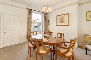 Une salle à manger avec une table ronde, six chaises, un lustre, une fenêtre avec des rideaux et des œuvres d’art encadrées accrochées aux murs.