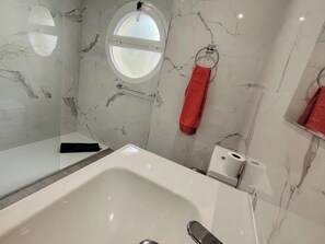 A bathroom with a large white bathtub, a round window, a towel ring with a red towel, and marble-patterned walls. - Piso de Lujo Cerca P. Banús - Vistas mar y montaña (Marbella)
