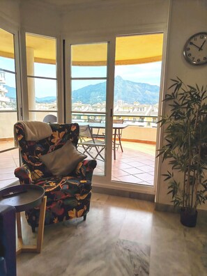 A room with a chair, a table, a clock, and a view of mountains through large windows. - Piso de Lujo Cerca P. Banús - Vistas mar y montaña (Marbella)