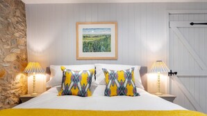 A bedroom with a stone wall, a framed landscape painting, two patterned pillows, a yellow bedspread, and two bedside lamps. - 3 Bed in Ambleside (oc-lr133) (Ambleside)