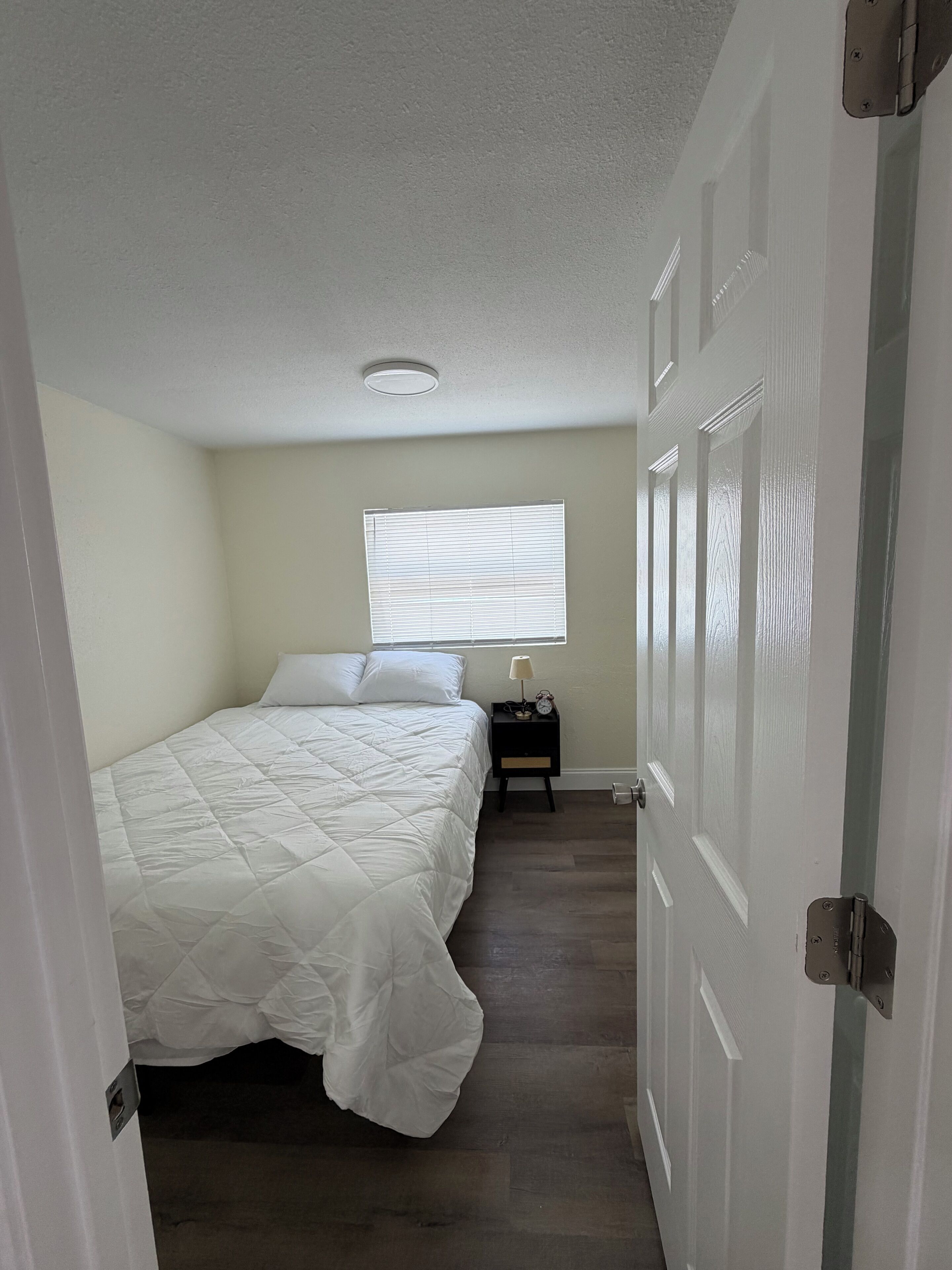 A bedroom with a bed, a nightstand, a window with blinds, and a door.