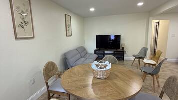 A dining area with a round wooden table, six chairs, a woven basket, a grey sofa, and a television in the background.