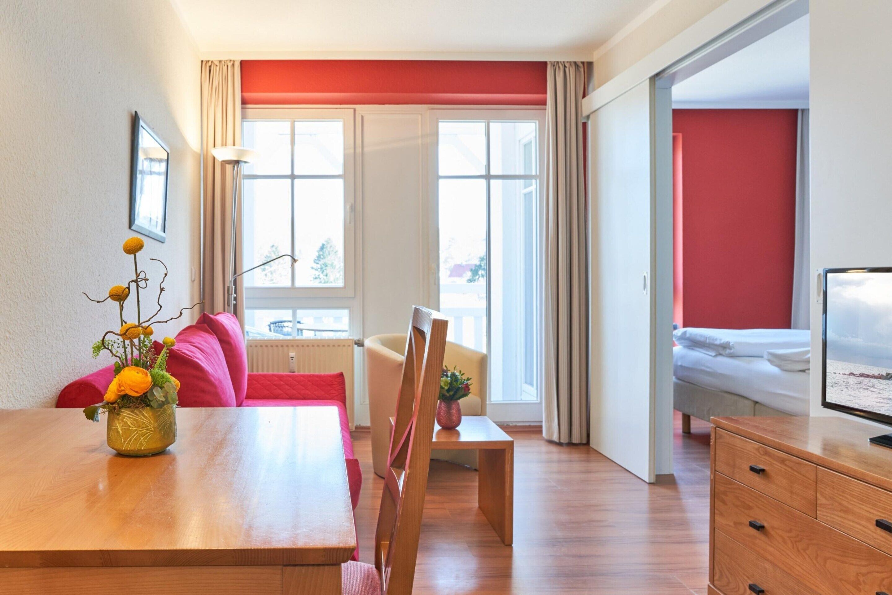 Ein modernes Hotelzimmer mit einem Holztisch, einer Couch, einem Sessel, einem Fernseher und einem Fenster mit Vorhängen.