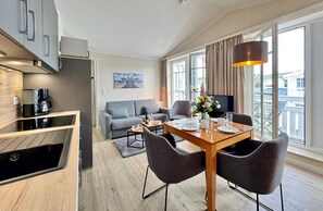 A modern kitchen with a built-in oven, a dining area with a wooden table and chairs, a grey sofa, and a large window with curtains. - Stylische Ferienwohnung mit Balkon und Wlan im Seepark Sellin (Sellin)