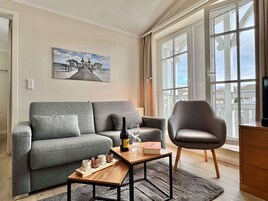 Un salon avec un canapé gris, une table basse en bois, un fauteuil en cuir, un tableau au mur et une grande fenêtre.