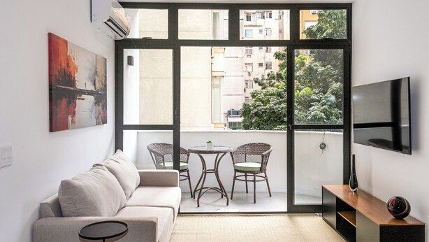 Ein modernes Wohnzimmer mit einer Couch, zwei Korbstühlen, einem kleinen Tisch, einem Fernseher und einem Balkon mit Blick auf die Stadt.