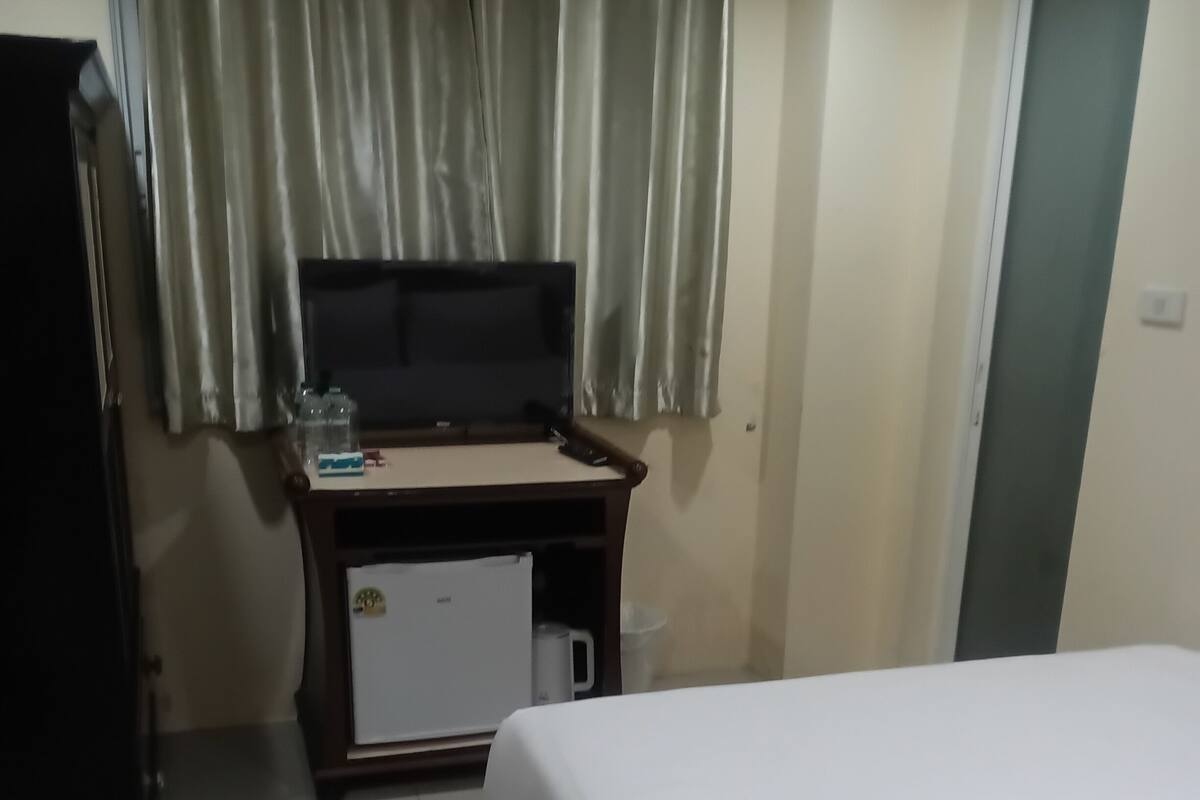 Ein Hotelzimmer mit einem Bett, einem Schreibtisch mit Computer, einer kleinen Kühlbox und einem Fenster mit Vorhängen.