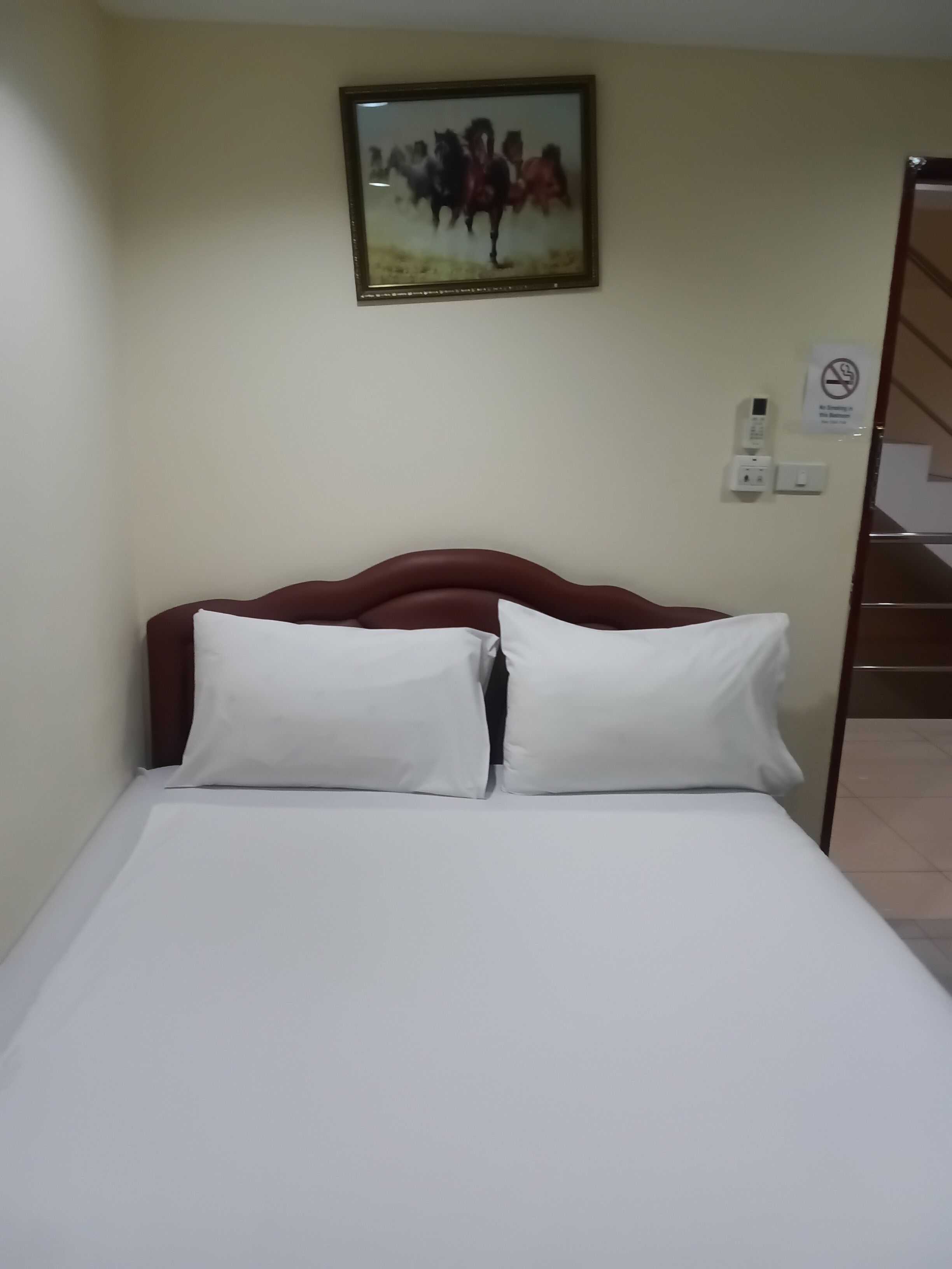 A hotel room with a bed, pillows, a wall-mounted painting, a telephone, and a no-smoking sign.