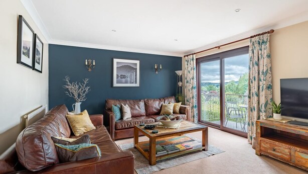 A living room with a brown leather sofa, a wooden coffee table, and a television on a wooden stand. - 4 Bed in Ambleside (oc-lr054) (Ambleside)