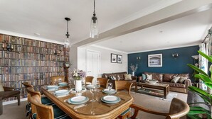 A dining area with a large wooden table set for six, surrounded by chairs with blue cushions. There is a bookshelf filled with books to the left, and a sofa with cushions in the background. - 4 Bed in Ambleside (oc-lr054) (Ambleside)