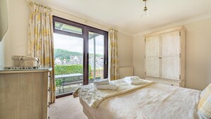 A bedroom with a bed, a wardrobe, a desk with a lamp, and a view of a green landscape through a large window. - 4 Bed in Ambleside (oc-lr054) (Ambleside)