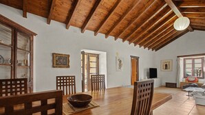Un comedor con mesa de madera, sillas y un tazón. Hay vigas de madera en el techo y un televisor en la pared.