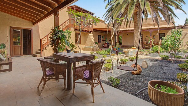 Un patio al aire libre con una mesa de madera y sillas de mimbre, rodeado de plantas en macetas y una palmera.