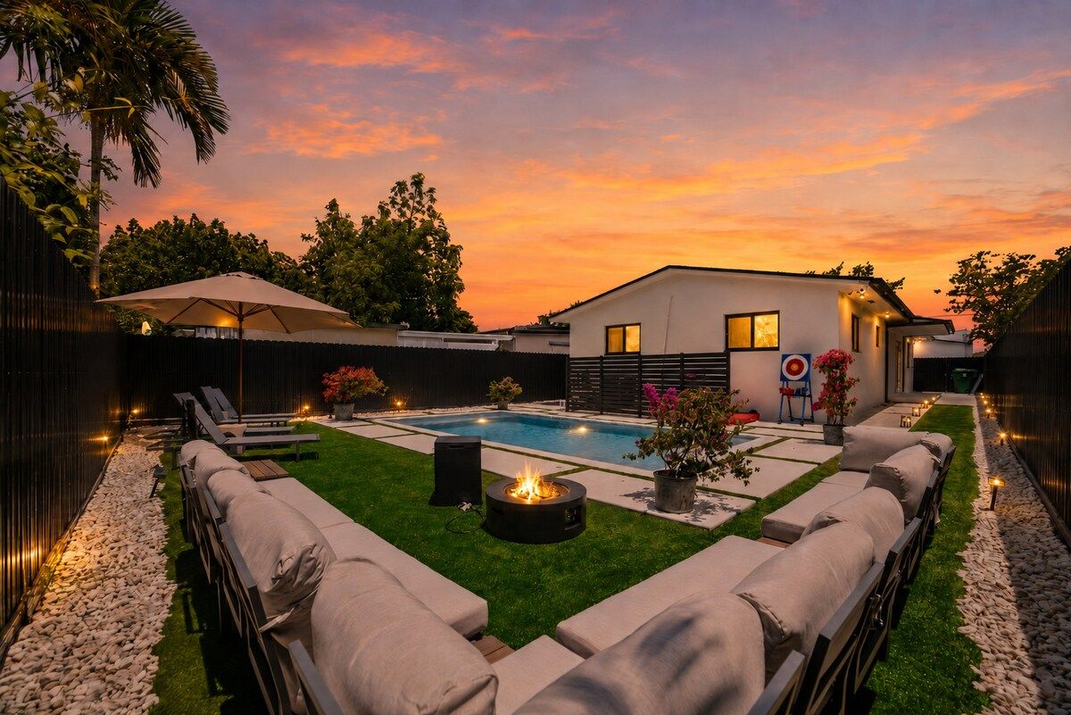 Un patio trasero con piscina, sillas de descanso, una fogata y una casa moderna.