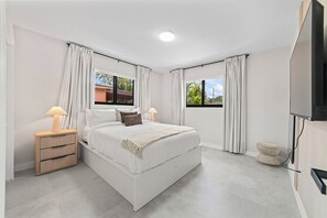 A modern bedroom with a large bed, two bedside tables with lamps, a TV, and a view of the outdoors. - Central Miami Home • Near Airport & Coral Gables (Miami)