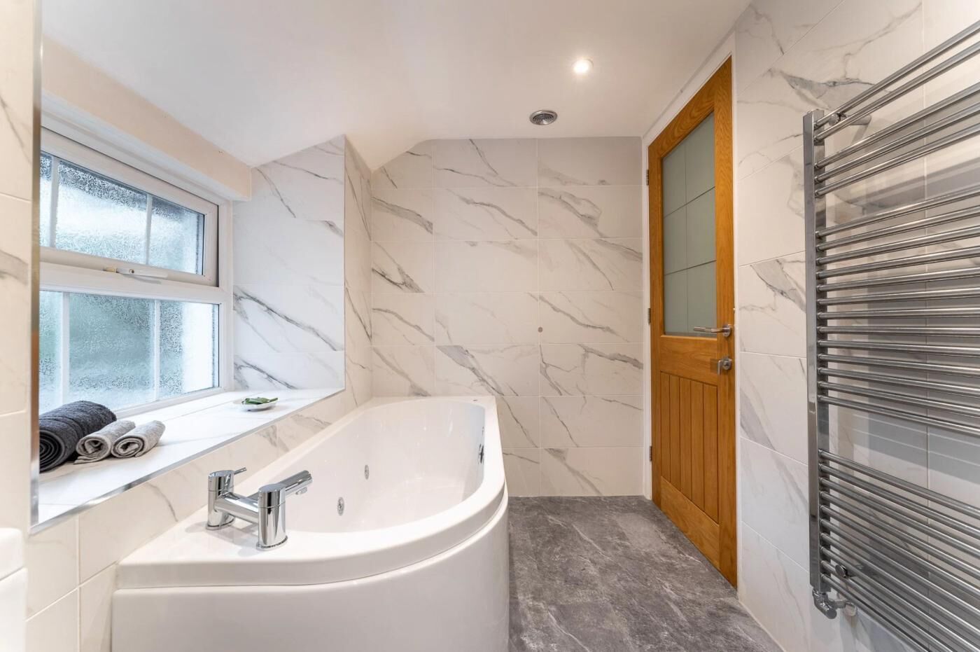 A modern bathroom with a freestanding bathtub, a window with frosted glass, and a wooden door.
