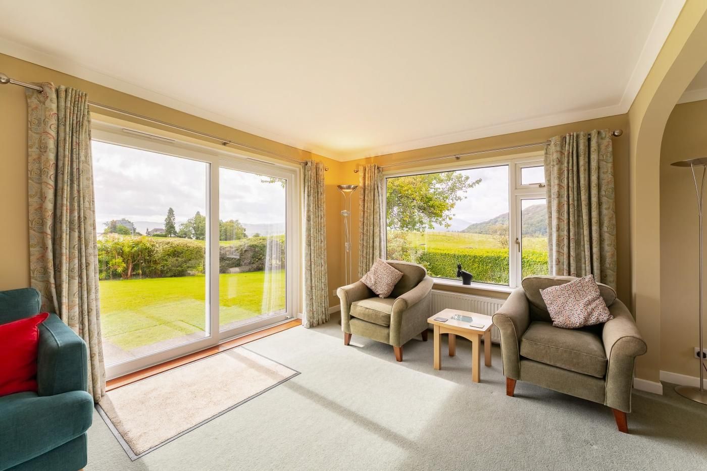 A spacious living room with a large sliding glass door leading to a garden, two armchairs, a small table, and a blue sofa.