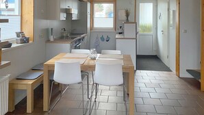 Et kompakt køkken med moderne hårde hvidevarer, et spiseområde med et træbord og hvide stole, samt udsigt gennem et vindue.