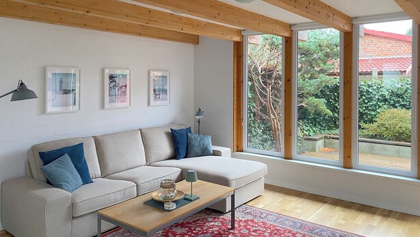 En moderne stue med sofa, sofabord og store vinduer med udsigt til grønne omgivelser.