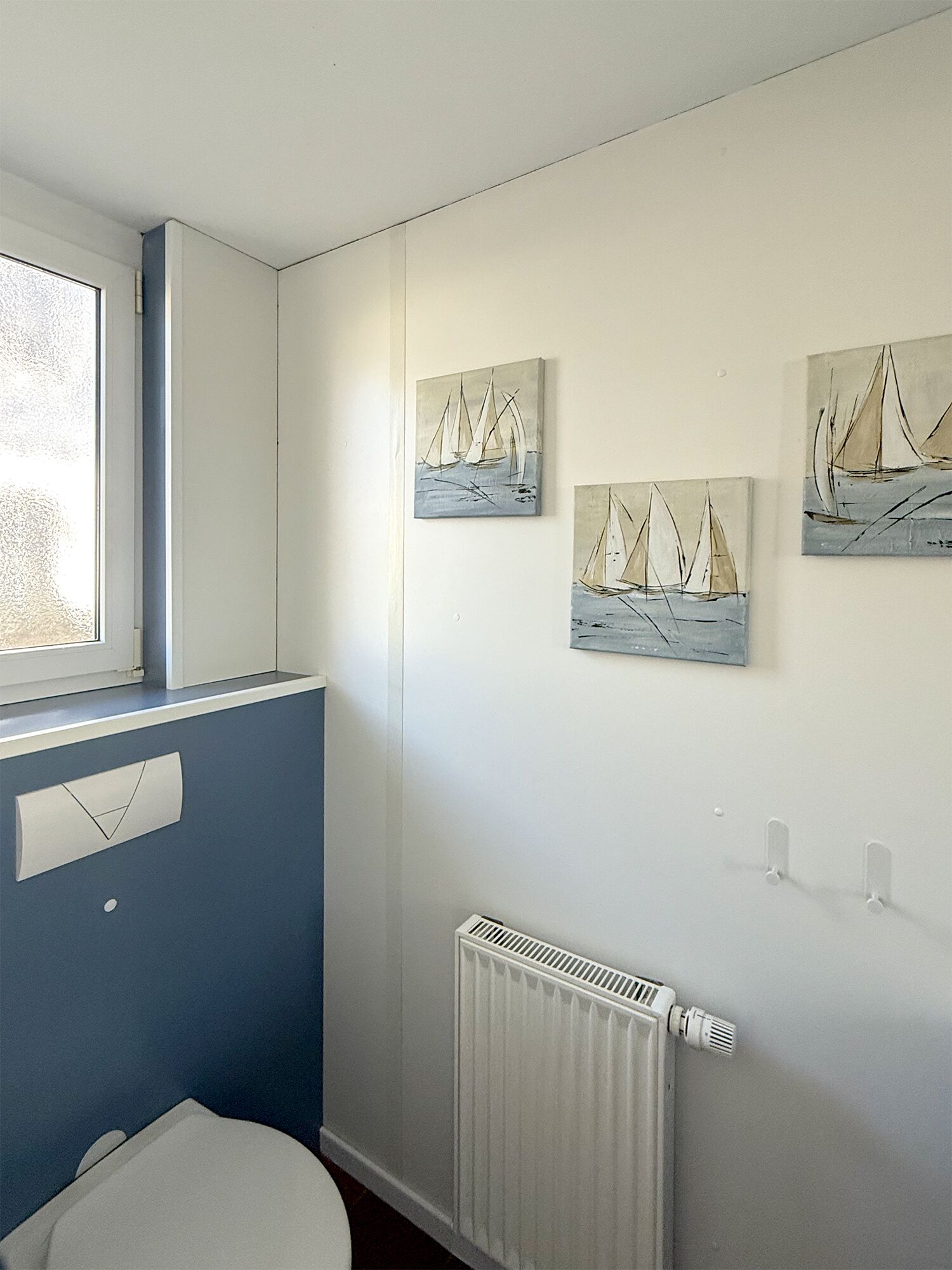 Une salle de bain avec des toilettes, un radiateur et trois tableaux représentant des voiliers accrochés au mur.