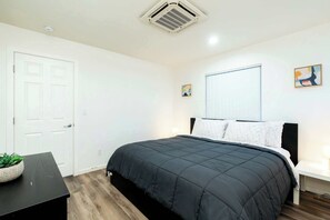 A bedroom with a black bedspread, white walls, a white door, and a window with blinds. - Foothills LA Family Friendly House (Glendora)
