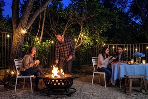 A group of people around a fire pit with a table and chairs, surrounded by trees and string lights. - Modern Cozy Stay w Outdoor Patio & Grill (Pomona)