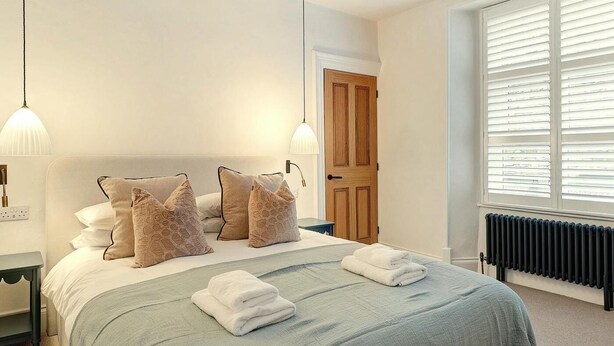 Una cama bien hecha con una colcha gris, dos almohadas y toallas dobladas. Se ve una puerta de madera y una ventana con persianas al fondo.