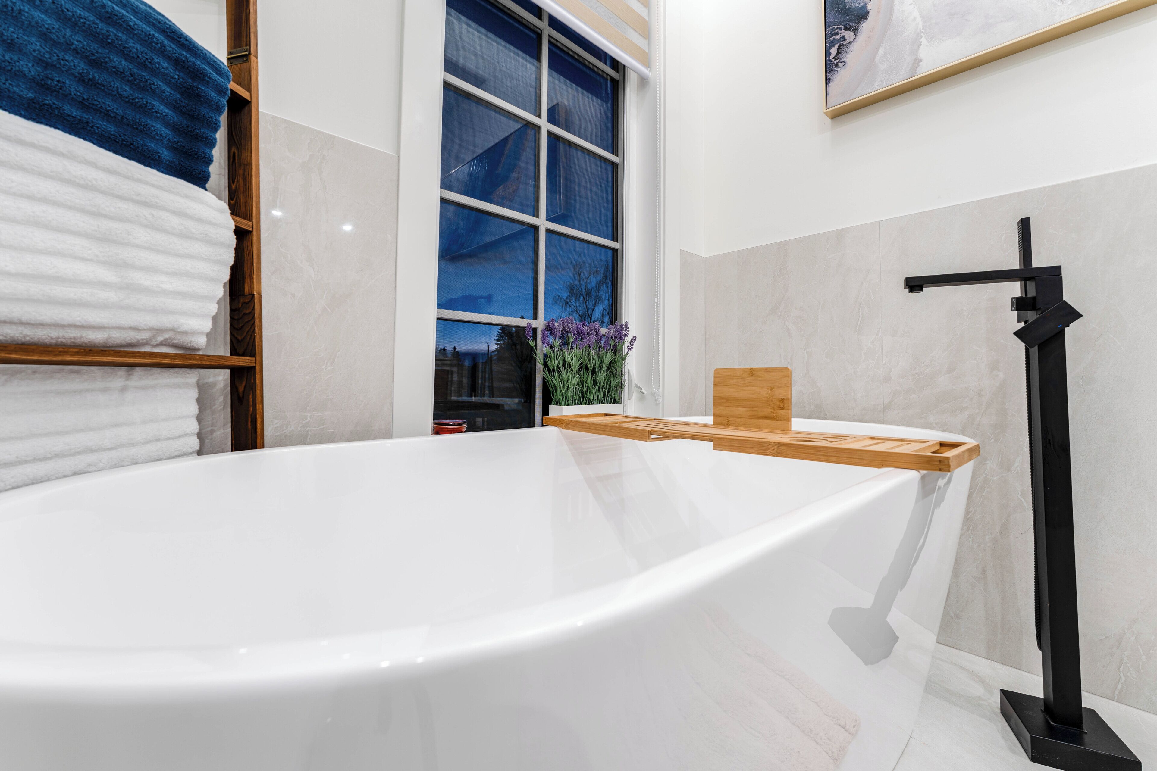 Un baño moderno con una bañera independiente, un estante de madera con toallas, un ventanal con vistas y un grifo negro.