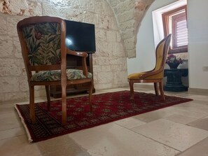 A room with a wooden chair and a television on a stand, a red patterned rug on the floor, and a stone wall. - Domus Clesia (Ostuni)