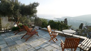 A patio with stone flooring, wooden chairs, and a table, overlooking a mountainous landscape.