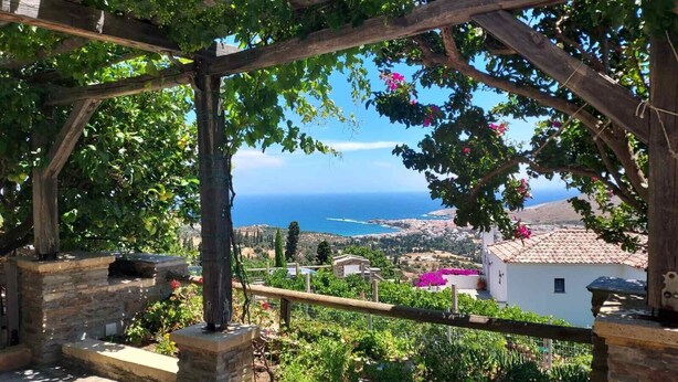 A stone-built structure with a wooden pergola, a stone bench, and a view of the sea and distant buildings.