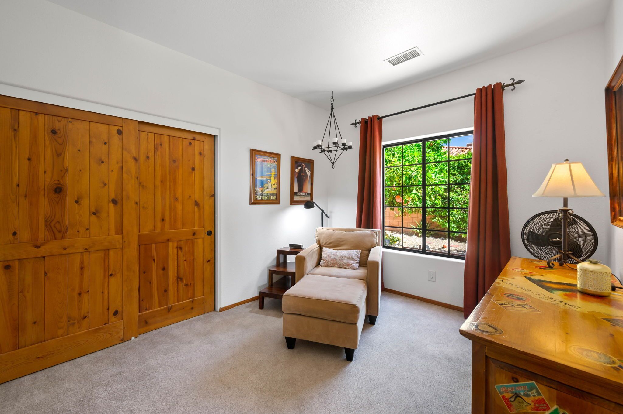 Una habitación con una puerta grande de madera, un sofá color beige, una mesita con una lámpara, un ventilador y una ventana con cortinas.