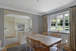 A dining area with a wooden table and chairs, a kitchen with white cabinets, and a window with curtains. - 3 Bed in Ambleside (oc-lr042) (Ambleside)